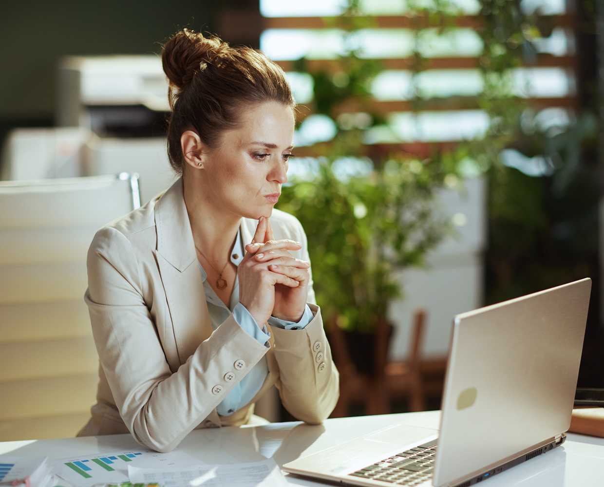 A woman in a blazer sits at a desk, looking at a laptop screen with a thoughtful expression, hands clasped under her chin. Papers with graphs are on the desk. Plants are in the background.