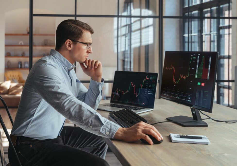 Man in glasses analyzes stock charts on two computer screens at a desk.